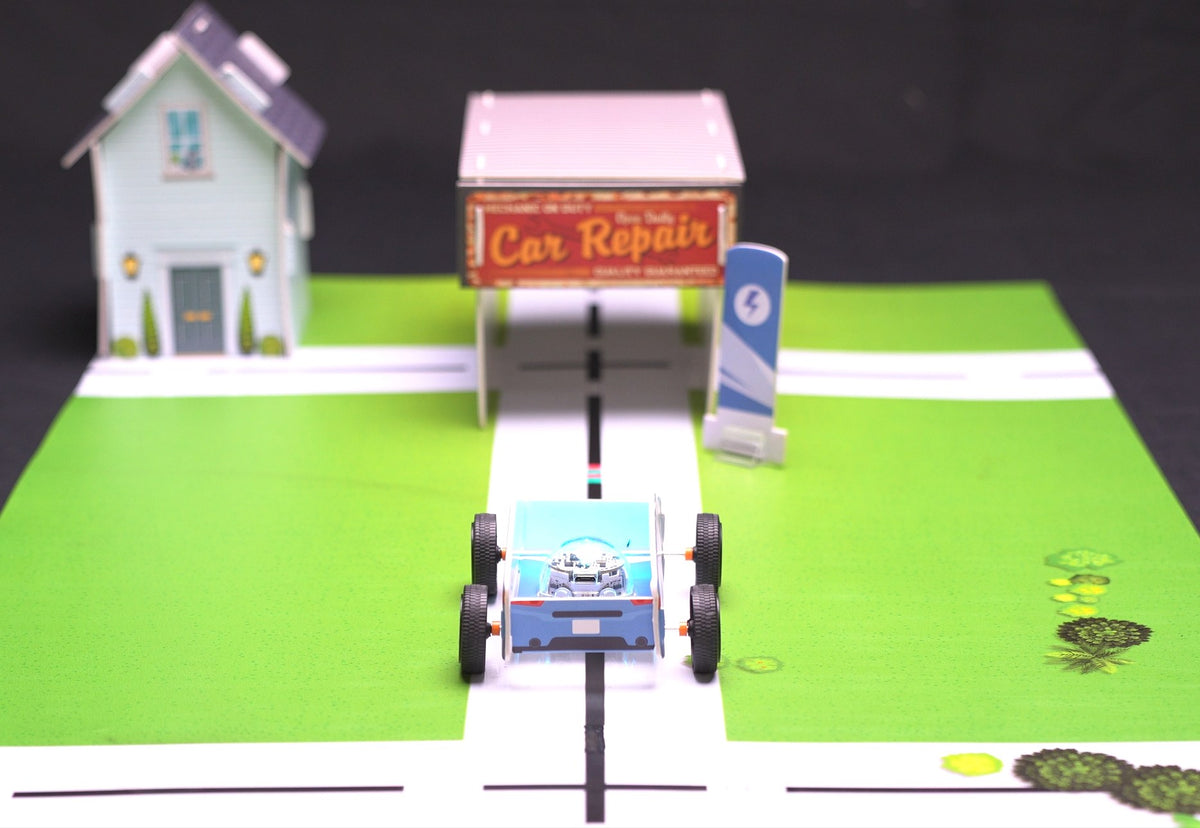 An Evo robot seated inside a cardboard car body, following a black line along a road on the neighborhood mat, with a paper house, a Car Repair garage and a charging post set up alongside.