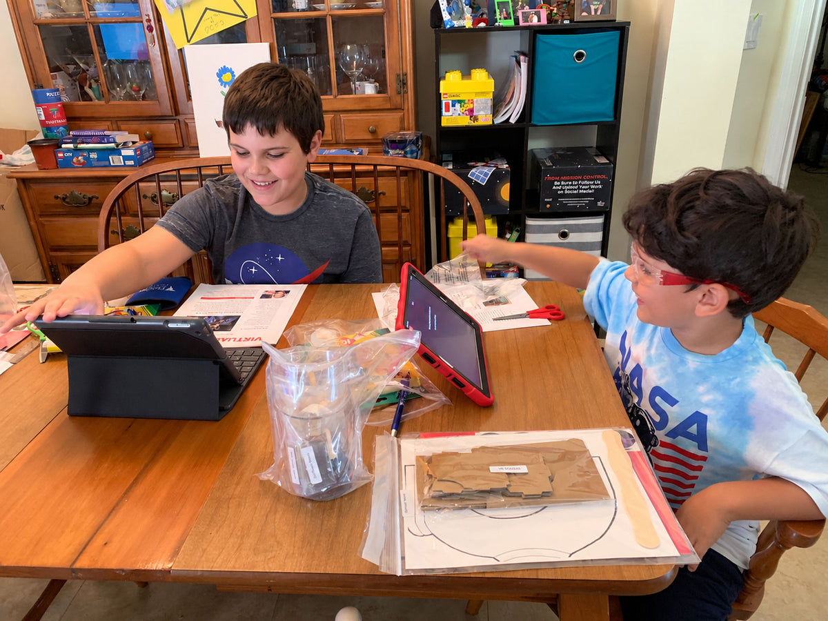 Two brothers in NASA shirts working the same activity at the family dining table, each with a tablet propped in front of them for the live session, the kit materials and the printed guide spread out between them.
