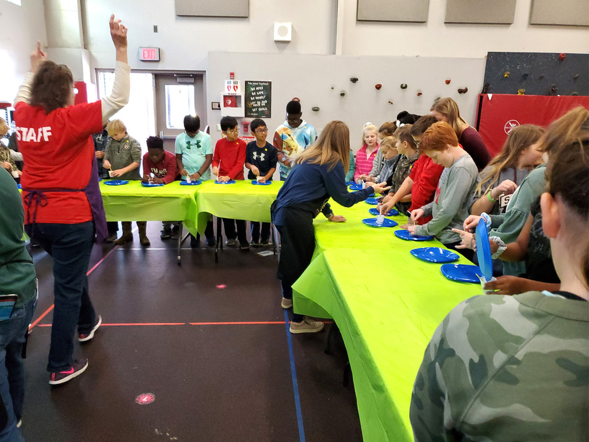 Students lined up along both sides of two long tables in a school gym, a plate of materials in front of each of them, while a Central Creativity staff member raises her arm to start the activity.
