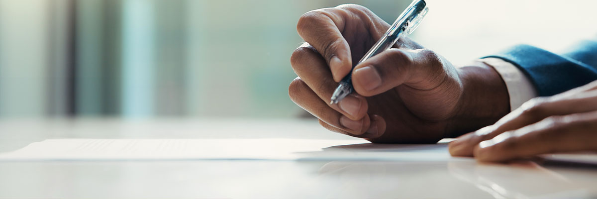 A close view of a hand signing a sheet of paper with a pen, the office behind it thrown out of focus.