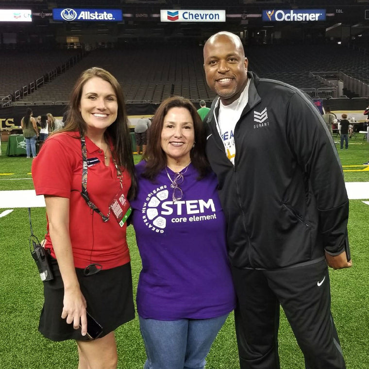 Jan Brenan, in her purple STEM Core Element shirt, stands arm in arm between two STEM Fest sponsors on the turf of the New Orleans Superdome, sponsor boards and empty stands stretching away behind them.