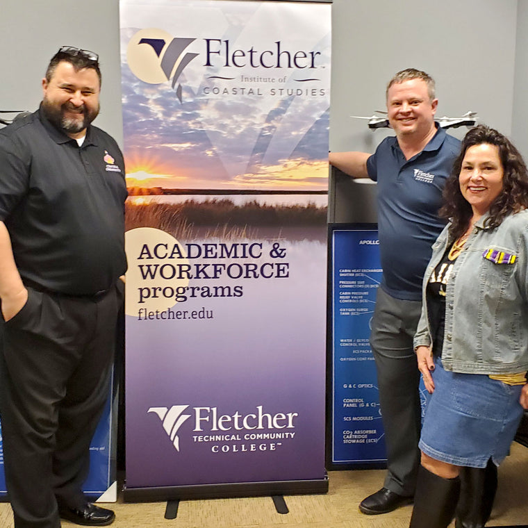 Jan Brenan stands at the right of a Fletcher Technical Community College banner beside Dr. Clint Coleman, the college’s Dean of STEM, with Rhye McLeod of Central Creativity in black at the left.