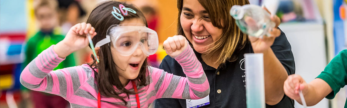 A young girl in oversized safety goggles raises both fists in triumph next to a grinning instructor holding up a bottle of foaming liquid.