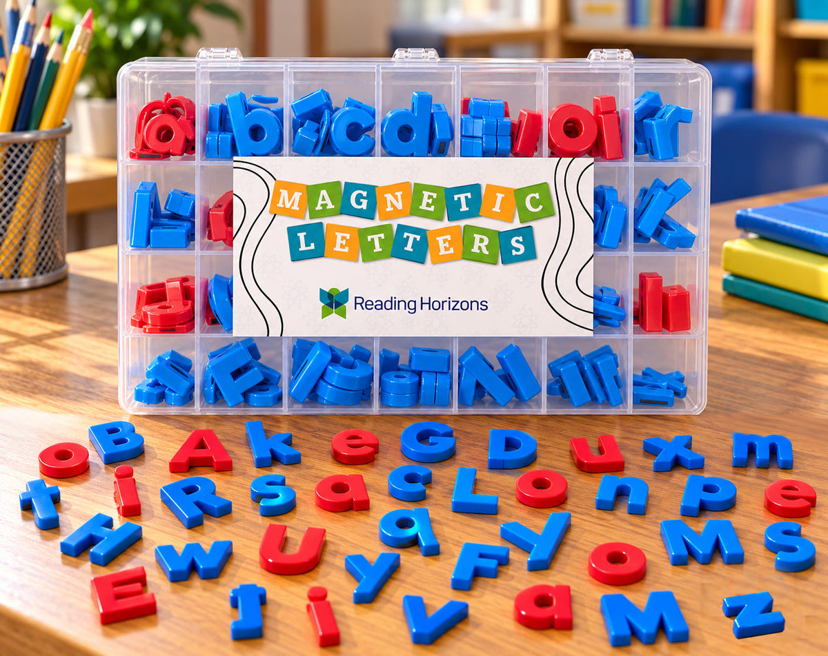 A clear compartment case of magnetic letters sits on a wooden classroom desk beside a cup of pencils and a stack of notebooks, with upper and lowercase letters spread out in front of it, the consonants in blue and the vowels in red.
