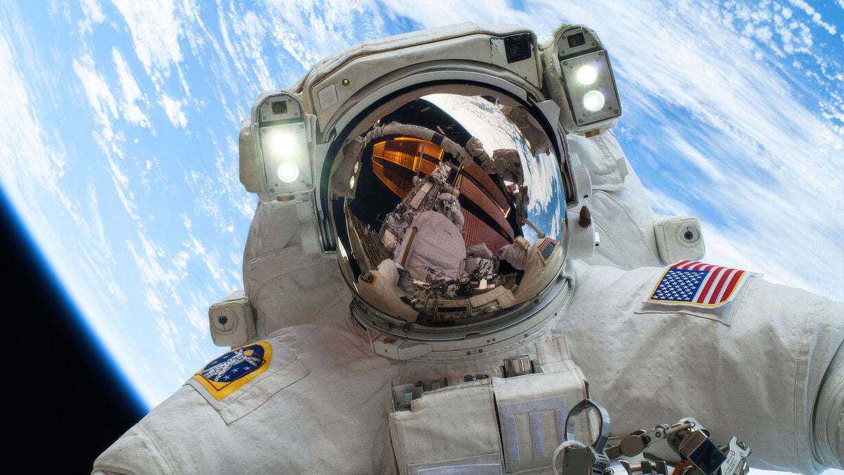 A NASA astronaut on a spacewalk high above Earth, the United States flag on the shoulder of the suit and the space station reflected in the helmet visor.