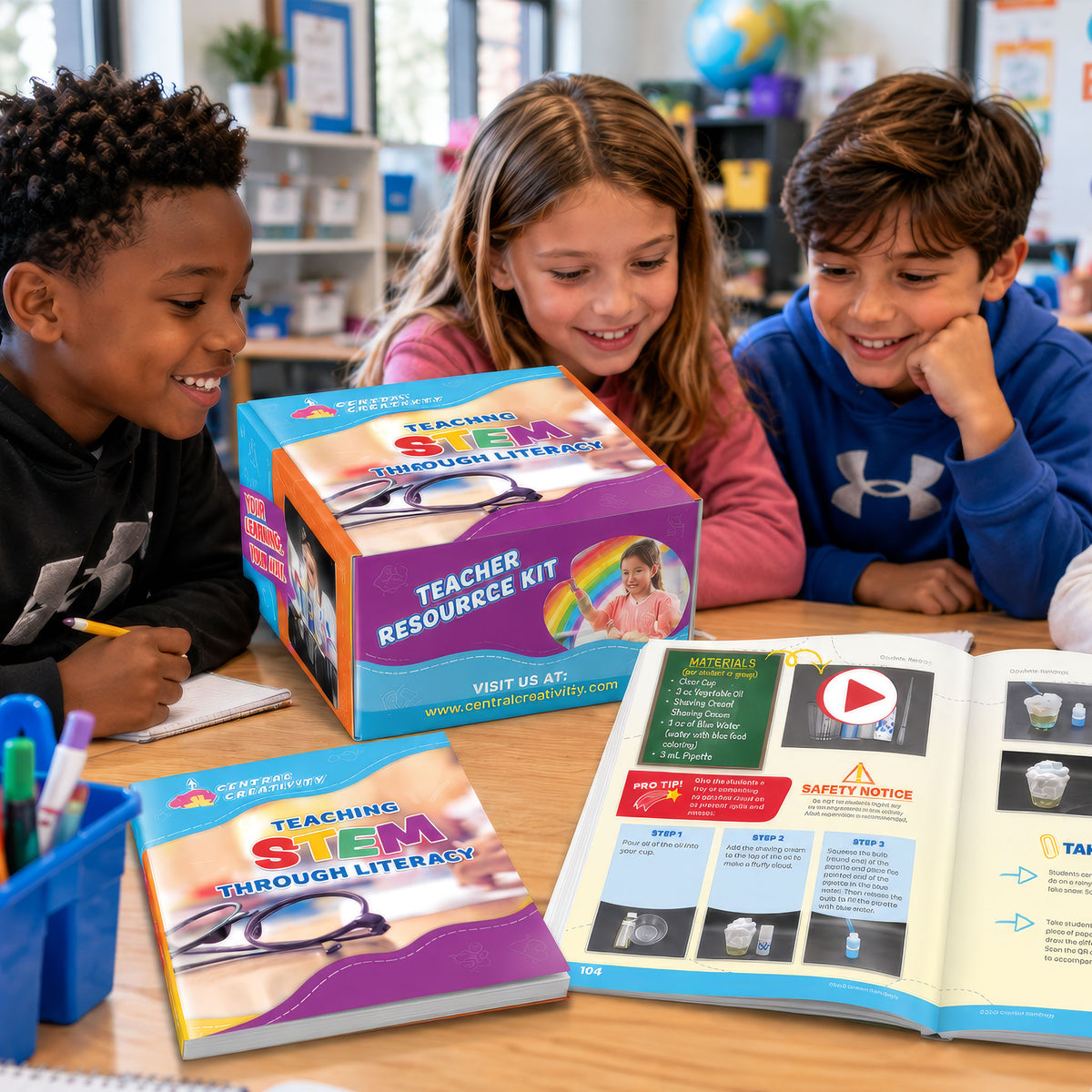 Three elementary students lean over a classroom table around the Teaching STEM Through Literacy Teacher Resource Kit box, with the teacher guide closed beside it and a second copy open to an illustrated activity spread.