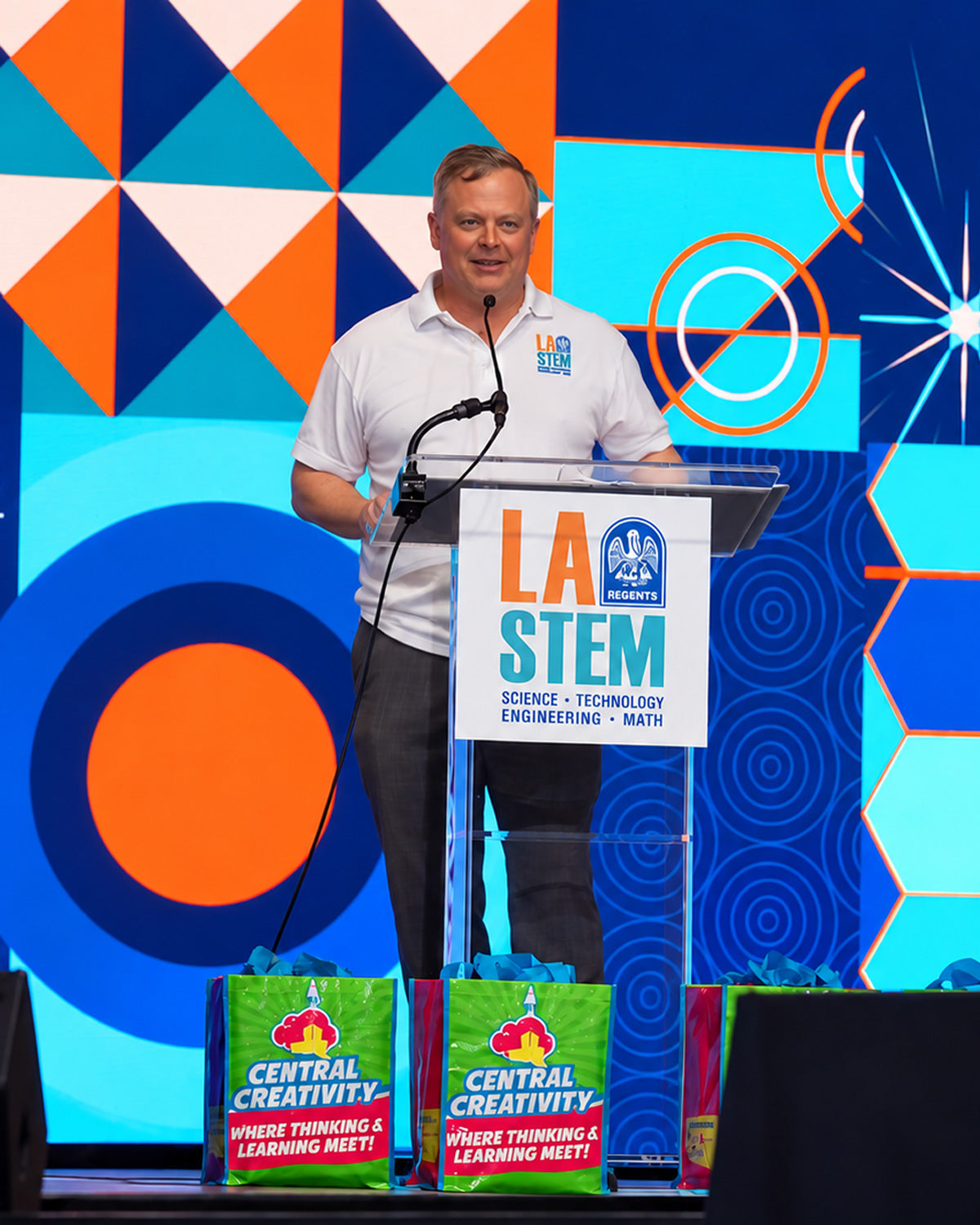 Dr. Clint Coleman speaking at a lectern branded LA STEM, in front of a bright blue and orange geometric backdrop, with Central Creativity kit boxes on the table beside him.