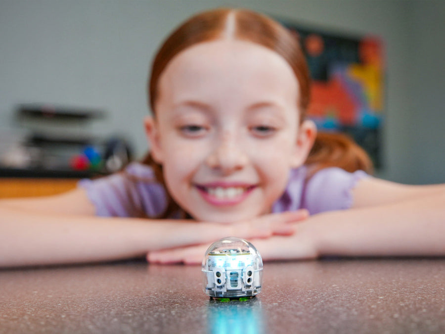 A girl leans on her folded arms at a table, smiling at a clear-domed Ozobot Evo robot lit white and teal a few inches from her face