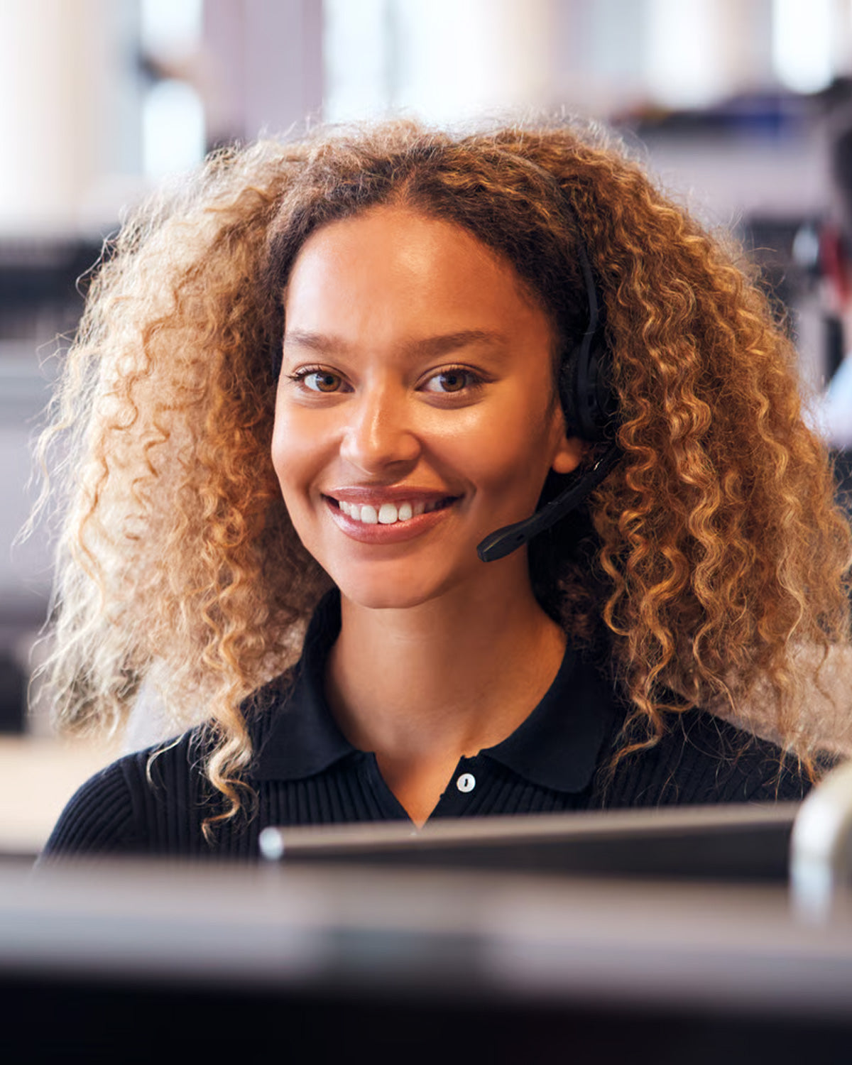 A support agent in a headset smiling at her monitor in an open-plan office.
