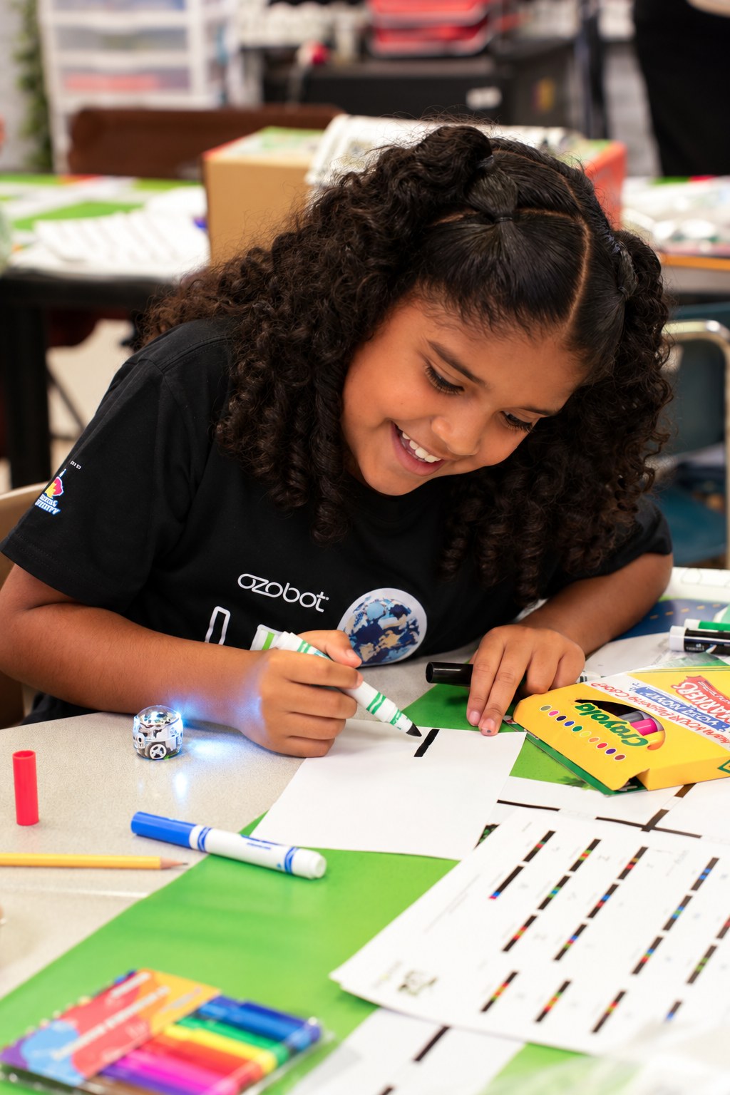 A student drawing a color code on paper with a green marker, an Evo robot lit up on the desk beside her.