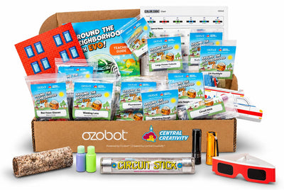 The Around the Neighborhood Teacher Resource Kit: a co-branded Ozobot and Central Creativity box holding a printed teacher guide, a Color Code chart, a cardboard building and labeled zip bags for each module, including Bee Vision Glasses, Welding Lens, Fluorescent, Modeling Dough, Large Flower Cutouts, Circuit Stick and UV Flashlight, with a cork tube, two small bottles, a Circuit Stick tube, two flashlights, red-framed glasses and a model vehicle laid out in front.