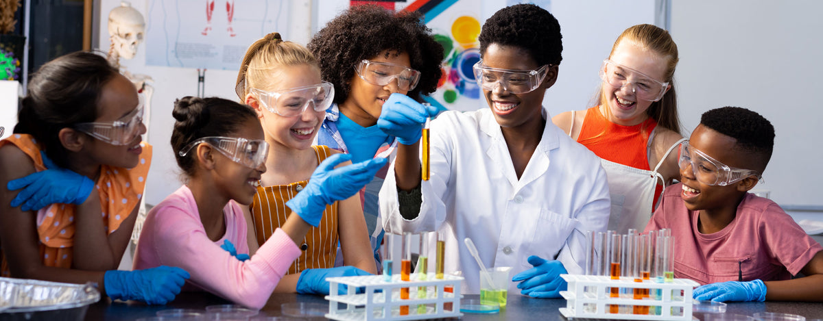 A teacher in a white lab coat and six students in safety goggles and blue gloves lean over a lab bench together, laughing as they fill racks of test tubes with colored liquid.