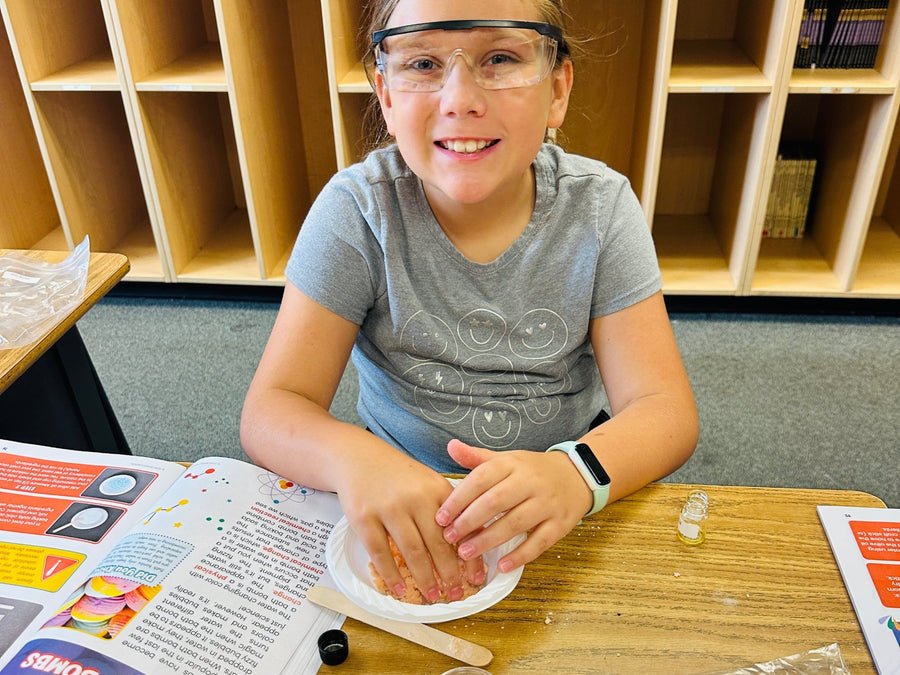 A girl in safety glasses pressing bath bomb mixture into a plate at her desk, with the guide open beside her.