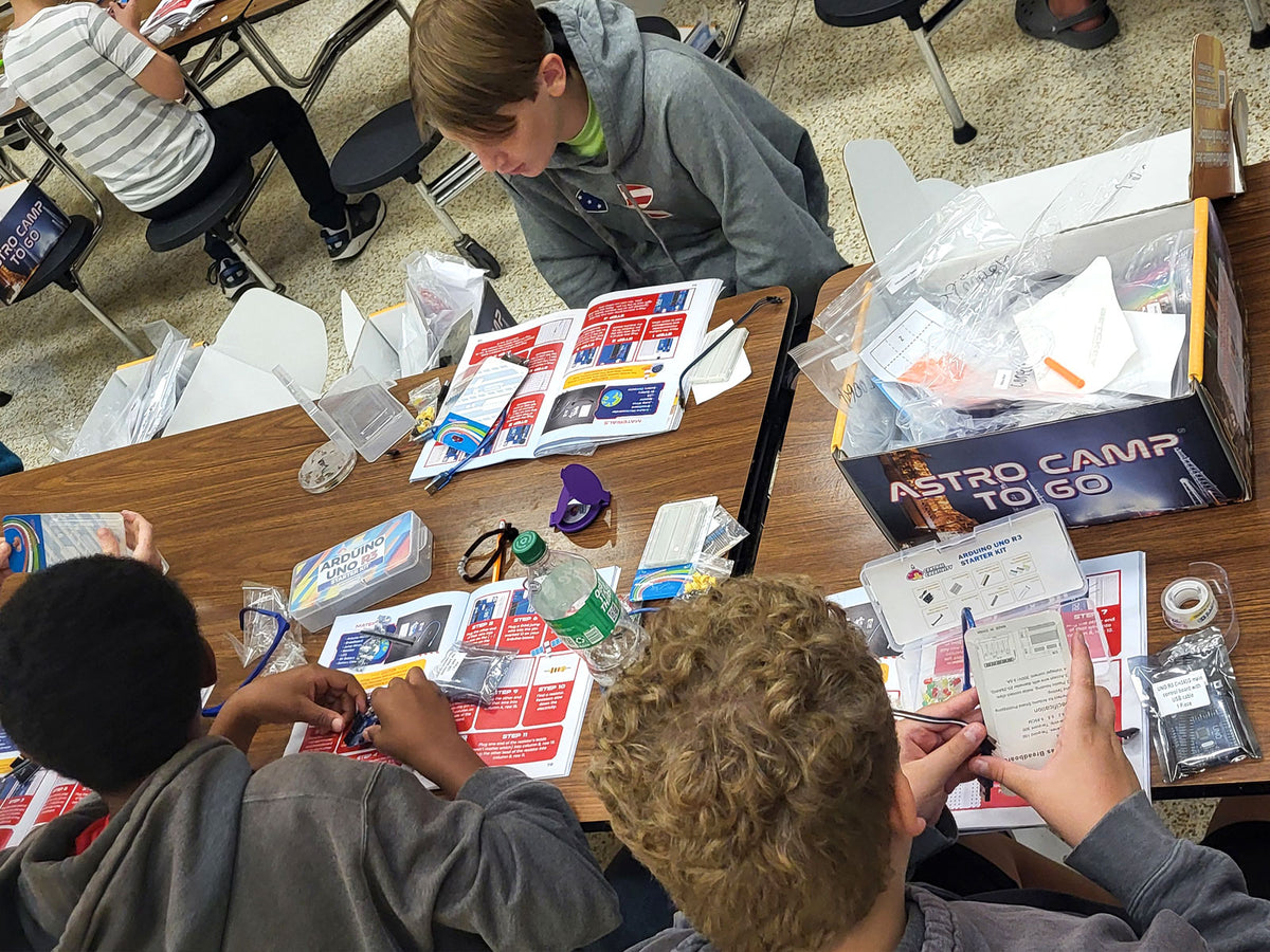 Looking down on a crew mid-session: four students around tables covered with bagged components and printed activity cards, an NASA ASTRO CAMP TO GO box open at the end of the table with its lid folded back.