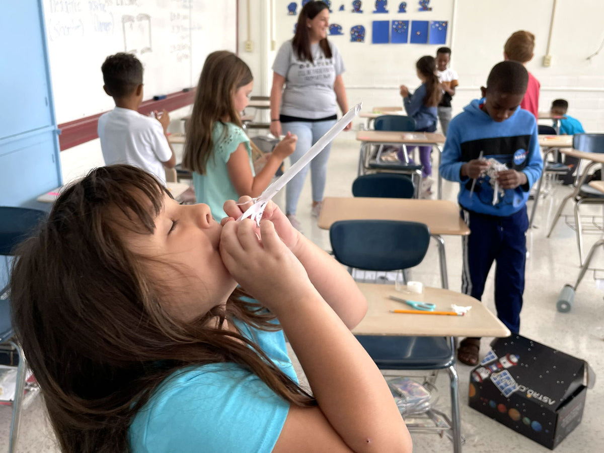 A girl tips her head back to launch a small paper rocket with a puff of breath, while behind her the rest of the crew works on their own builds and an adult moves between the desks rather than standing at the front.