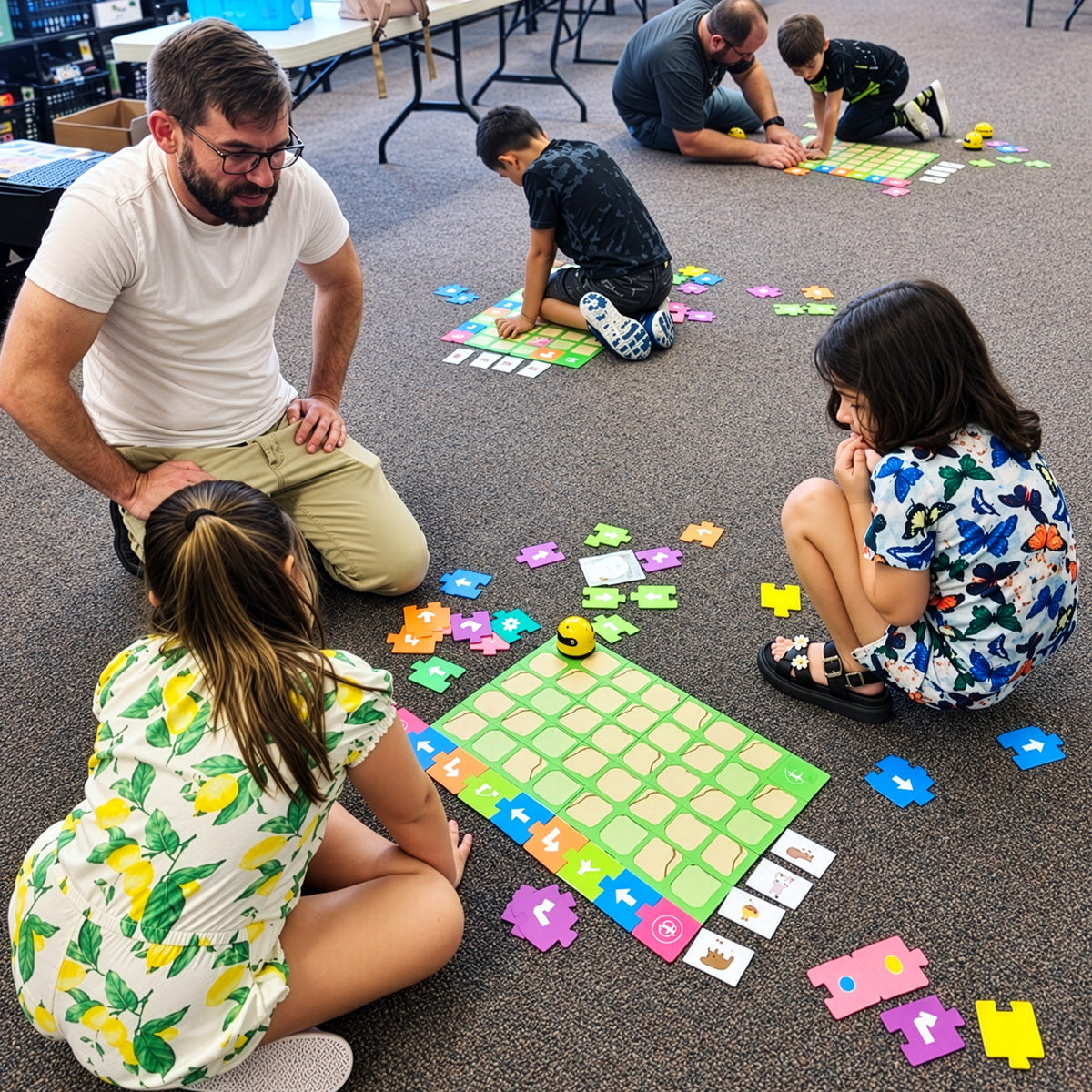 Children sitting on a classroom carpet around a green grid mat, placing colored arrow tiles to program a small yellow robot while two adults kneel alongside them