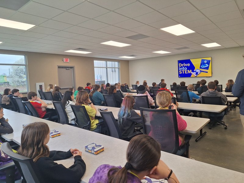Students seated facing a screen reading Drive Into STEM, each with an unopened kit box on the table