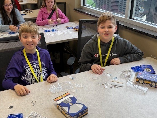 Two students smiling at a table with kit boxes and loose parts in front of them