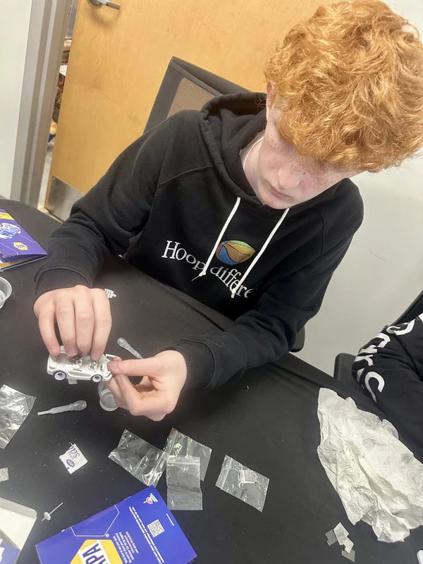 A student fitting the chassis of his Saltwater Car together, with part bags, a pipette and a NAPA instruction card on the table in front of him.
