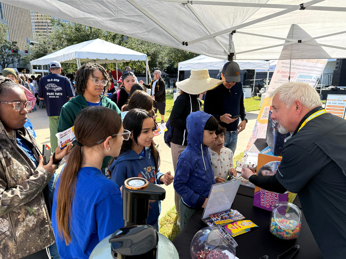 Children crowd a Central Creativity table at an outdoor STEM festival while a staff member in a lanyard hands out an activity under a white event tent