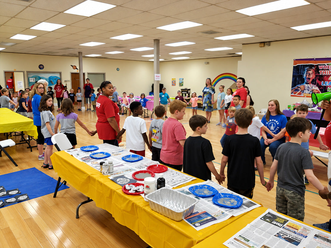 A long table under a yellow cloth laid out with plates, foil trays and newspaper, a line of children waiting along one side of it, and more children and staff in red shirts at tables across the hall behind.