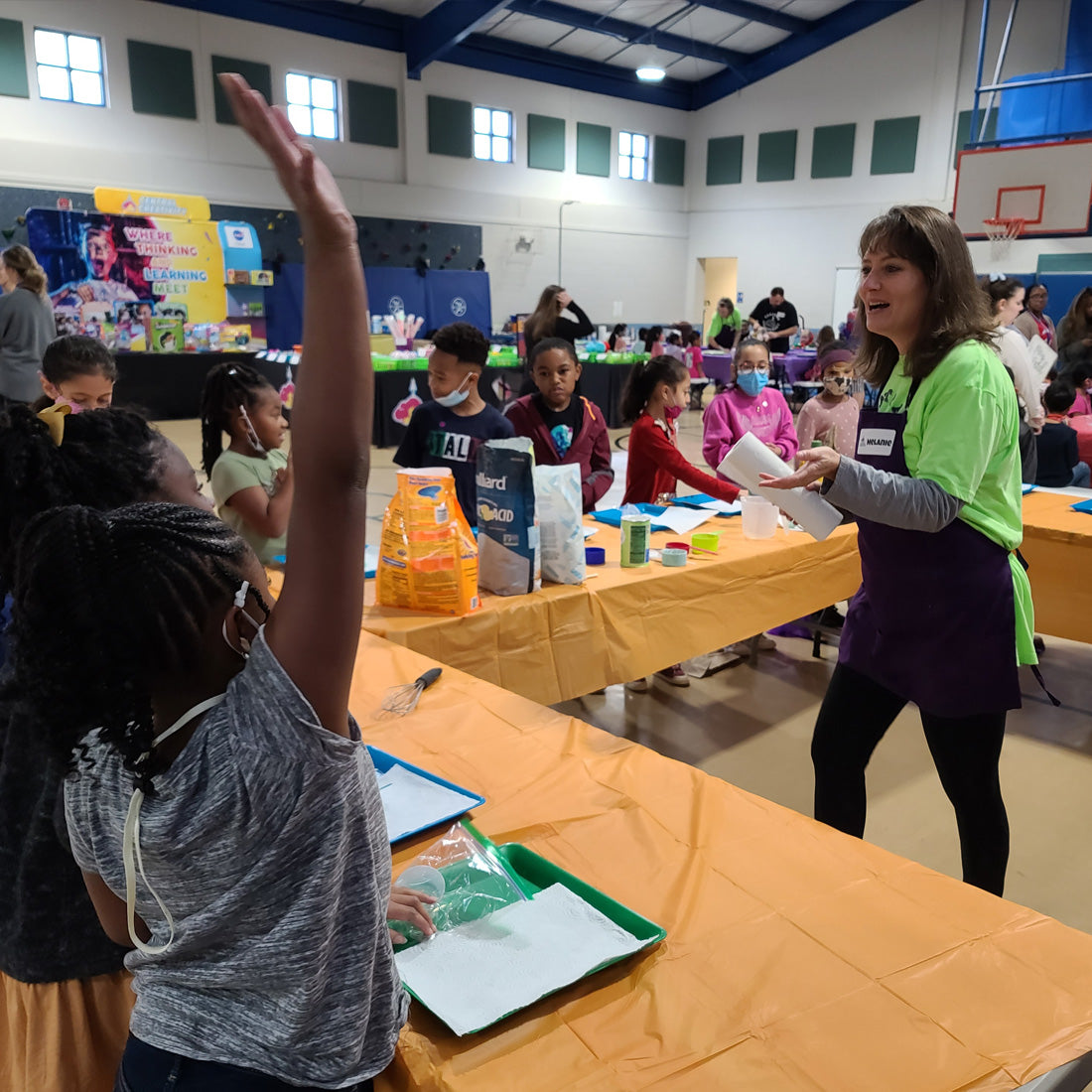 A student at a long activity table raises her hand high while a Central Creativity facilitator in a bright green shirt turns toward her, with more students working at tables down the length of a school gym behind them.