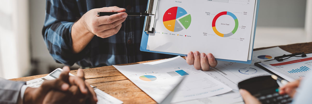 A hand holding a pen points at a clipboard of pie and donut charts, with printed reports spread across the table around it.