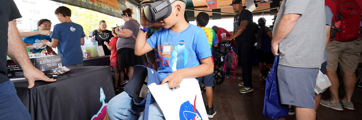 A boy tries a virtual reality headset at an outdoor community event while staff in matching shirts run activity tables and families move between them.