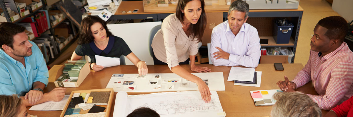 Six colleagues around a conference table covered with plans and drawings, one of them standing and leaning in to point something out.
