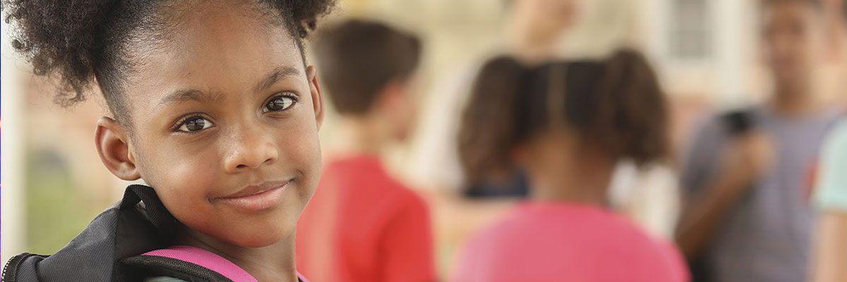 A young girl in a classroom smiles toward the camera, other children at work behind her out of focus.