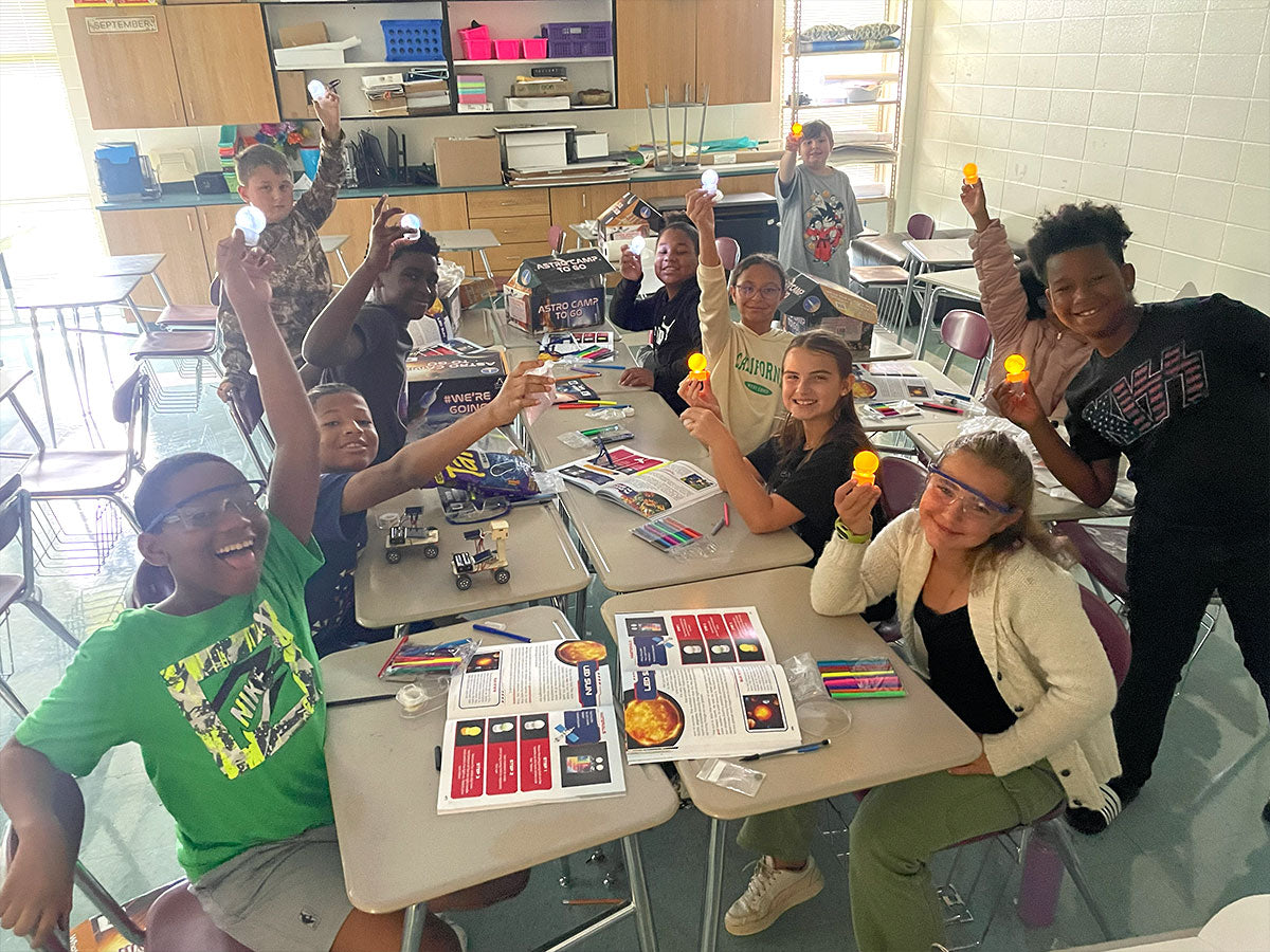 Eight or nine children crowd around classroom tables, every one of them holding a small yellow canister up toward the camera and grinning, with printed activity guides and a robotics kit spread out in front of them.