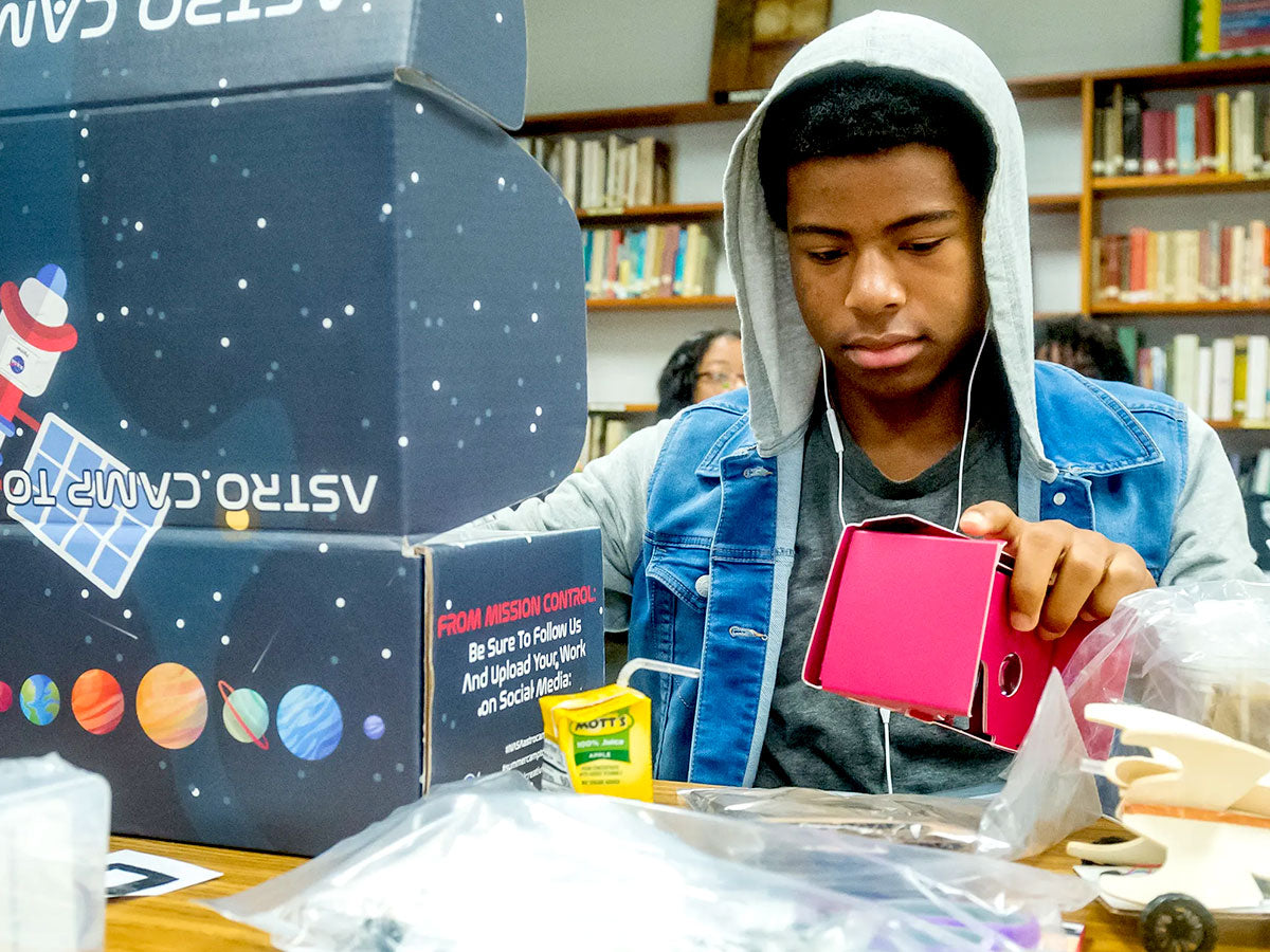 A teenager in a hooded sweatshirt works intently on a bright pink component at a library table, an NASA ASTRO CAMP TO GO kit box printed with planets open beside him and parts laid out in bags.