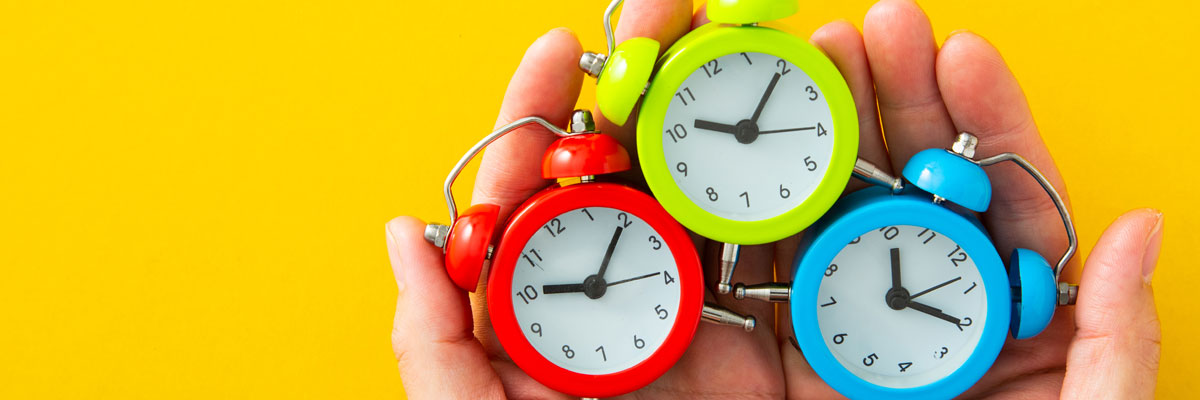 A pair of cupped hands holding three small alarm clocks &mdash; red, green and blue &mdash; against a bright yellow background.
