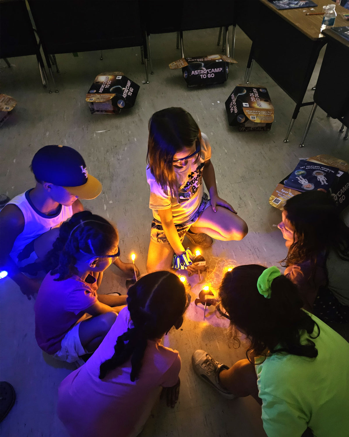 Six children kneeling in a circle on a classroom floor in low light, watching small glowing lights and a trail of smoke between them, with NASA ASTRO CAMP To Go kit boxes stacked on the tables behind them.