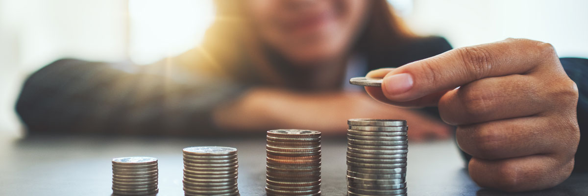 Four stacks of coins rising step by step across a table while a smiling person out of focus behind them adds one more coin to the tallest stack.