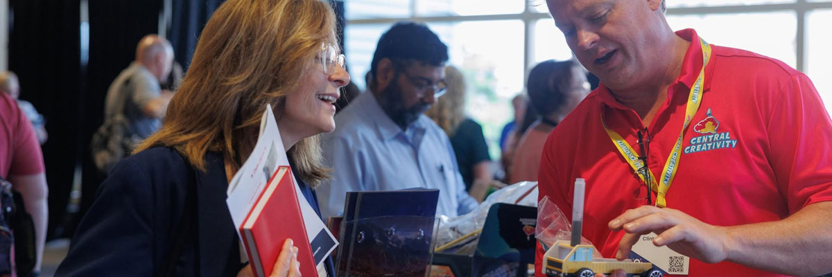 A man in a red Central Creativity polo shows a small yellow model truck to an educator holding a folder of handouts, across a busy exhibit table with other visitors crowded behind them.