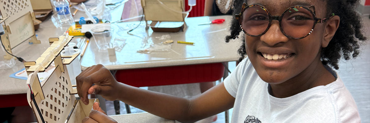 A girl in tortoiseshell glasses grins back over her shoulder while her hands keep working on a laser-cut plywood build, at a classroom table covered with kit parts, tools and other students&rsquo; projects.
