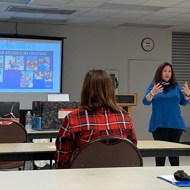 Jan Brenan, in a blue top, speaks with her hands to a seated room of professors and STEM leaders at Nicholls State University, beside a projected slide headed Region 3 and filled with photographs from across the network.