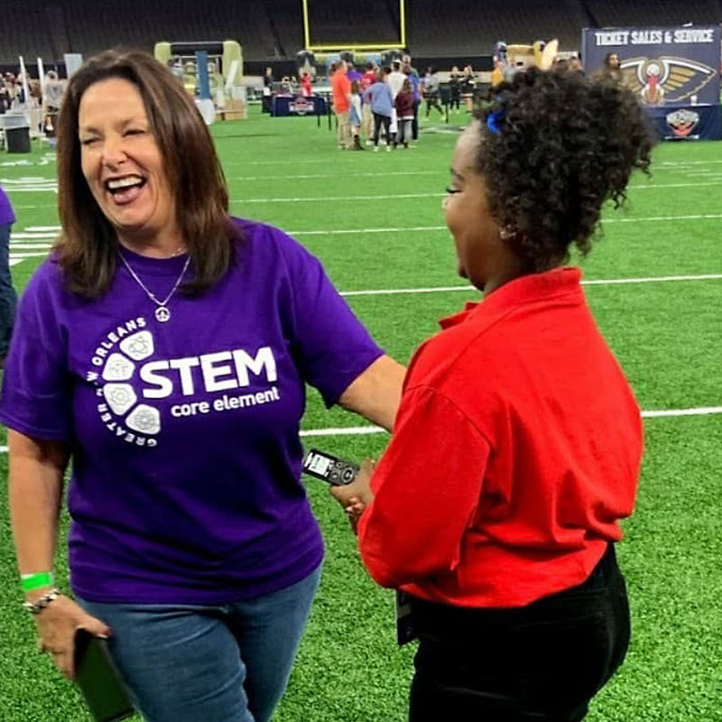 Jan Brenan, laughing in her purple STEM Core Element shirt, being interviewed on the field at STEM Fest in the New Orleans Superdome.
