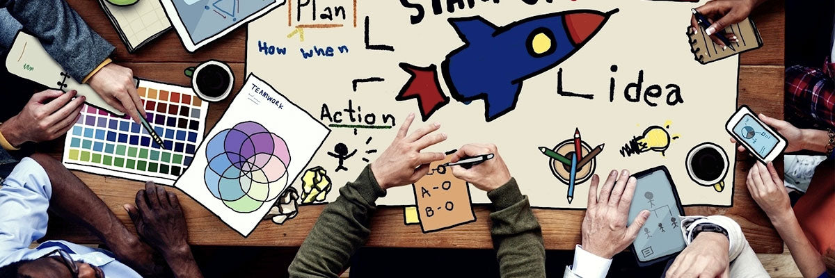 An overhead view of a table where several people work together on a large hand-drawn plan marked Idea, Plan and Action around a rocket, with color swatches and notes spread around it.