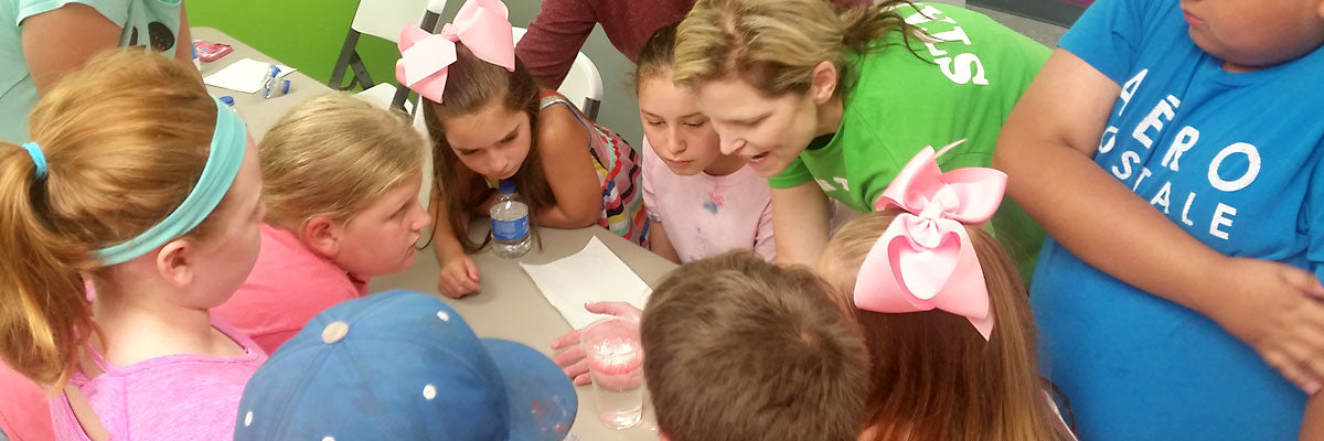 A cluster of young children leans in over an activity table together, heads almost touching, watching closely as an adult helper guides what is happening in front of them.