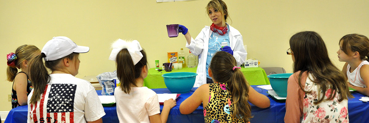 A volunteer in a lab coat and gloves demonstrates an experiment at an activity table while a group of children lean in to watch.