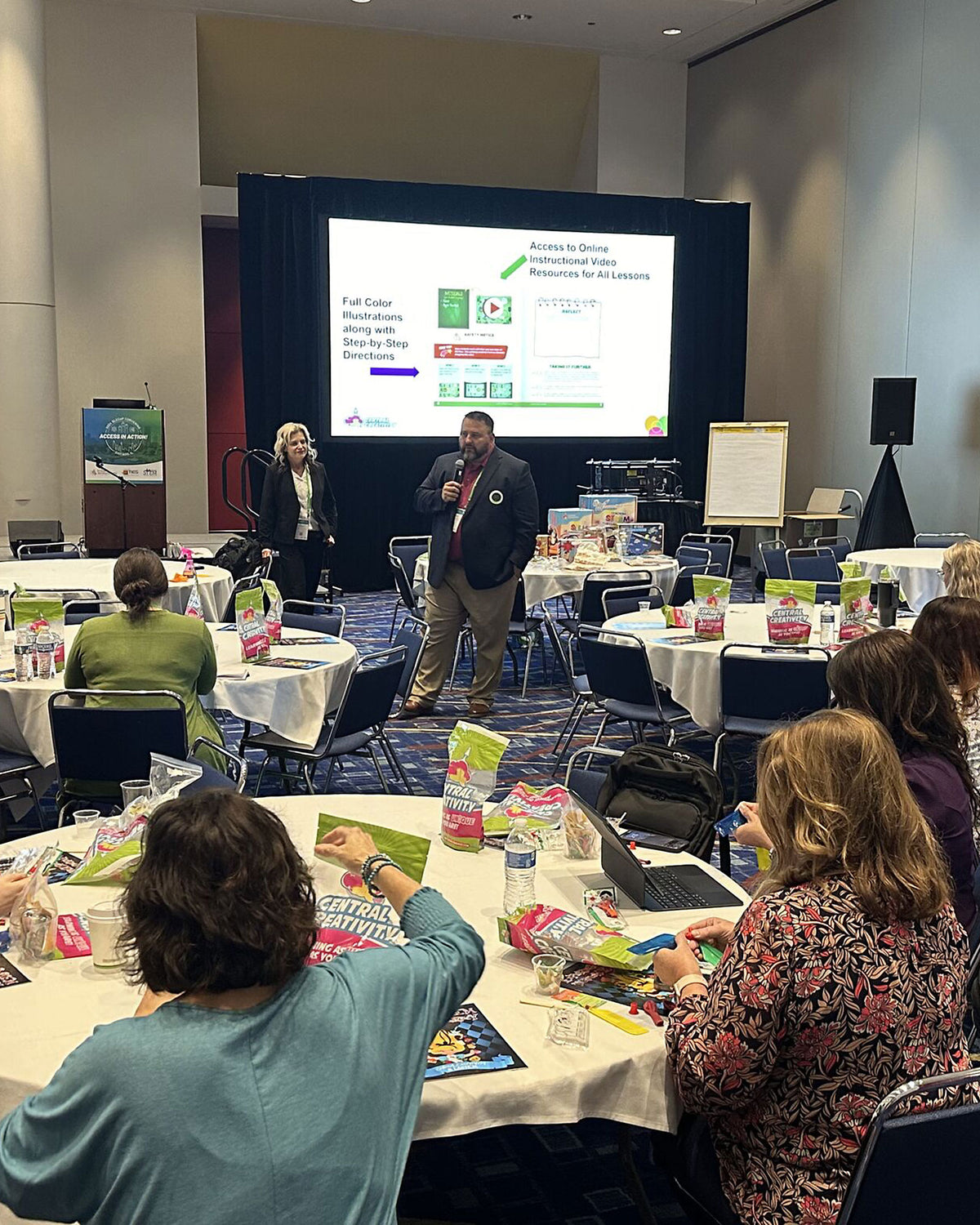 A presenter speaks into a microphone at the front
        of a ballroom while attendees at round banquet tables work through Central Creativity kit
        materials, a slide behind him listing the illustrated step-by-step directions and online
        video resources included with every lesson.