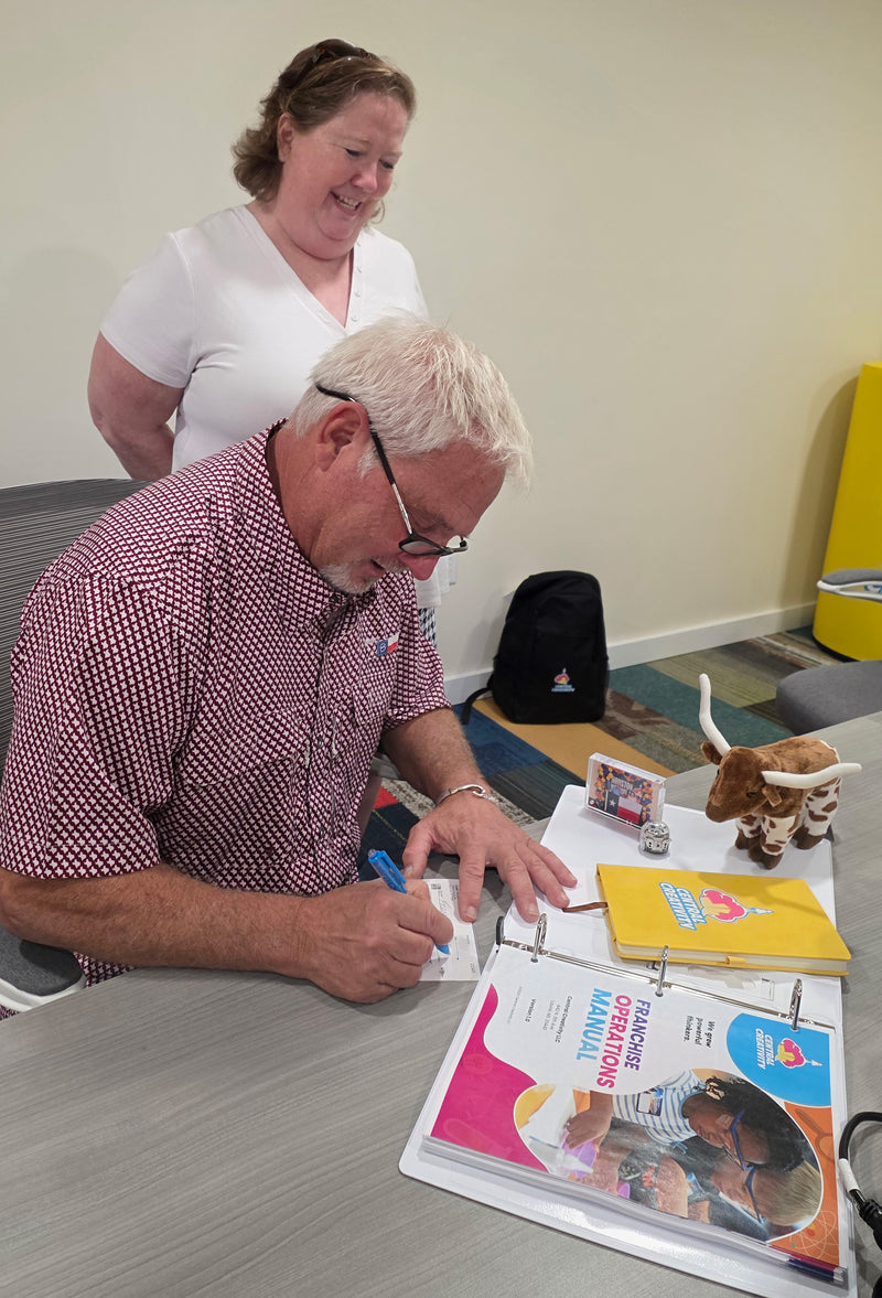 A new franchise owner leaning over a table signing the Central Creativity franchise operations manual, a colleague standing beside him.