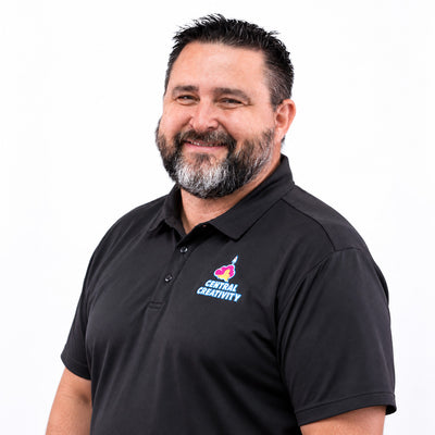 Roger “Rhye” McLeod, smiling with his arms folded, wearing a black Central Creativity polo shirt against a plain light background.