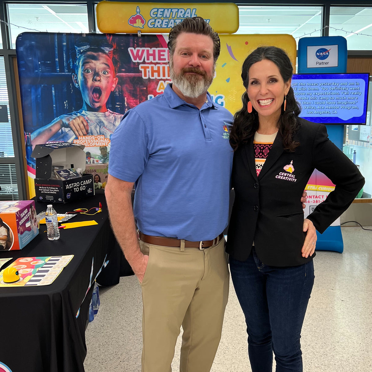 Kerry and Chris Wahl standing arm in arm and smiling in front of a Central Creativity trade show display, Kerry wearing a branded jacket.