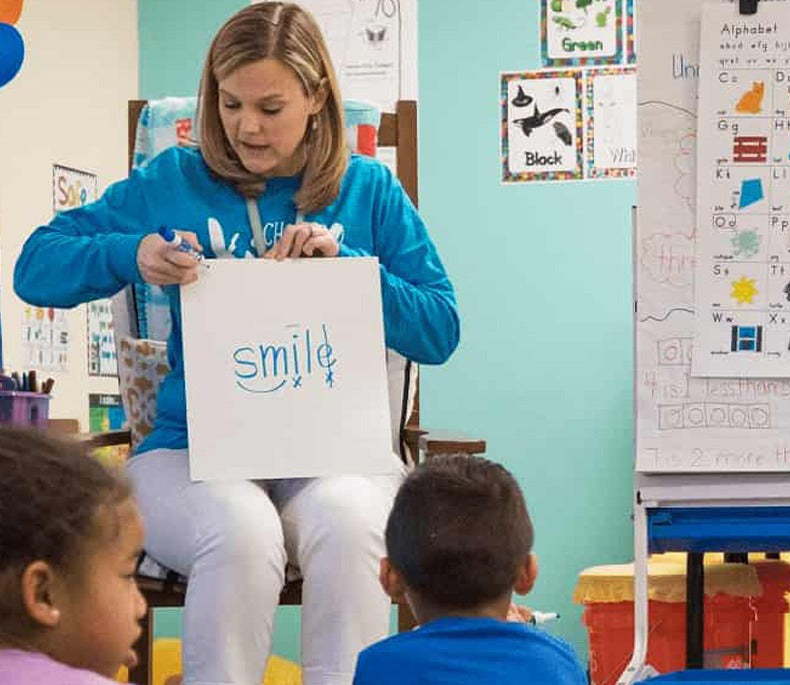 A Reading Horizons teacher holding up a card with the word smile marked for decoding, a bar over the i and a line through the silent e, while children sitting on the floor write on their own boards.