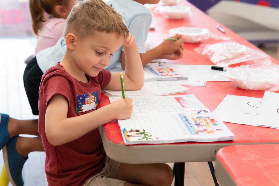 A boy in a red shirt writing with a pencil in an open Hands-On Literacy student guide at a long table, a Hands-On Literacy name badge on his shirt, with other children working their way along the table beside him.