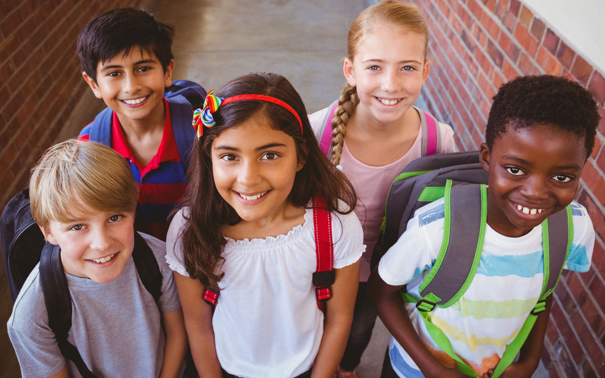 Five elementary school students wearing backpacks stand together outside a brick school building, looking up at the camera and smiling.