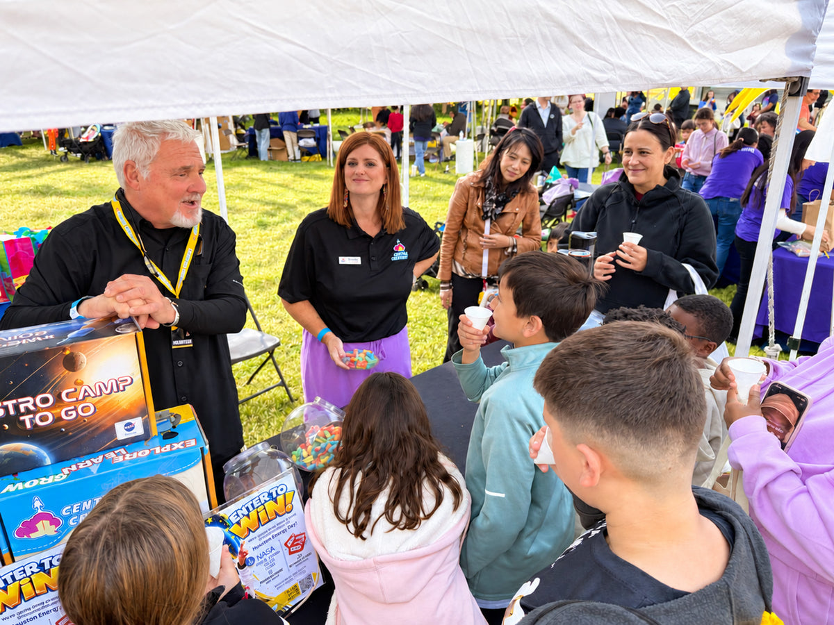 Children crowd a Central Creativity tent at Houston Energy Day, sipping from small paper cups while staff hand out more beside stacked ASTRO CAMP TO GO kits